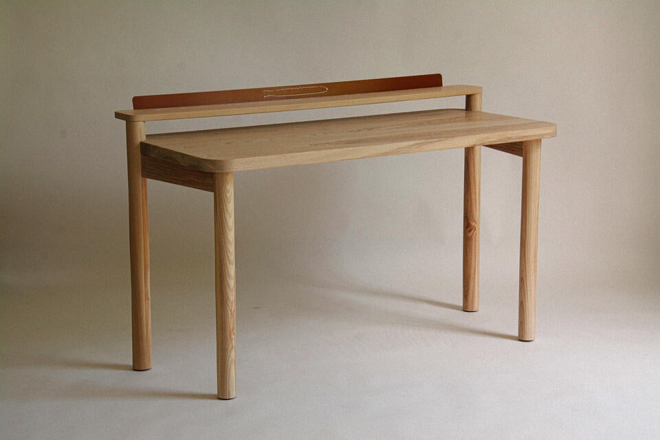 A solid ash desk of standard proportions, with an upper ledge made out of steel to be used to store postcards or assorted ephemera. 