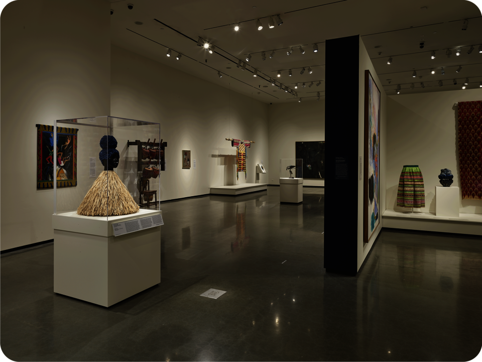 Installation view of an exhibit. A partial wall with a black painted side extends towards the viewer. Visible are colorful textile works, paintings, ceramics, and figurative sculptures. 