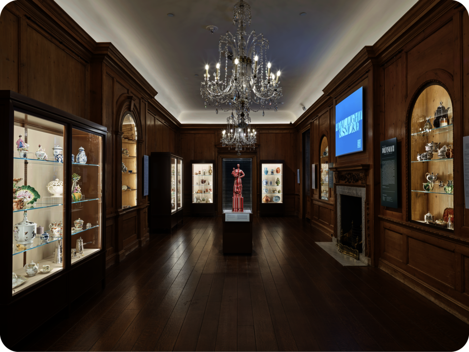 Exhibit documentation. Cases containing ornate ceramics and metalwork line the three walls of the space. A striped statue stands in the space’s center below a large chandelier. 