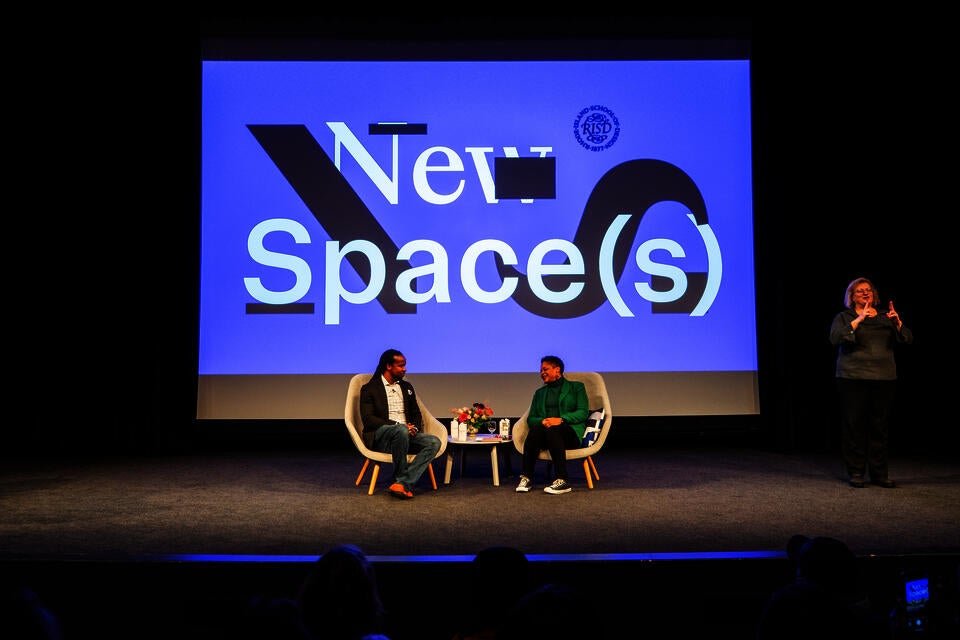 A Black man and woman sit on a stage, conversing. The New Space(s) logo is projected behind them, and an ASL interpreter stands to the side.