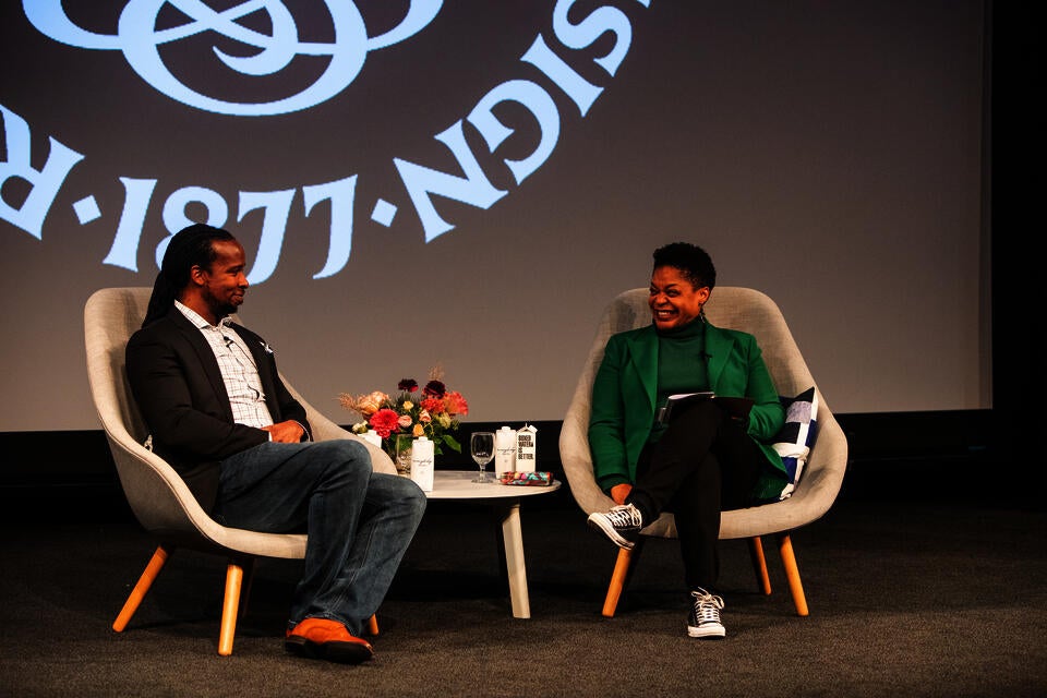 A smiling Black man and woman sit on a stage. Between them is a table with flowers and boxes of water. The RISD seal is projected behind them.