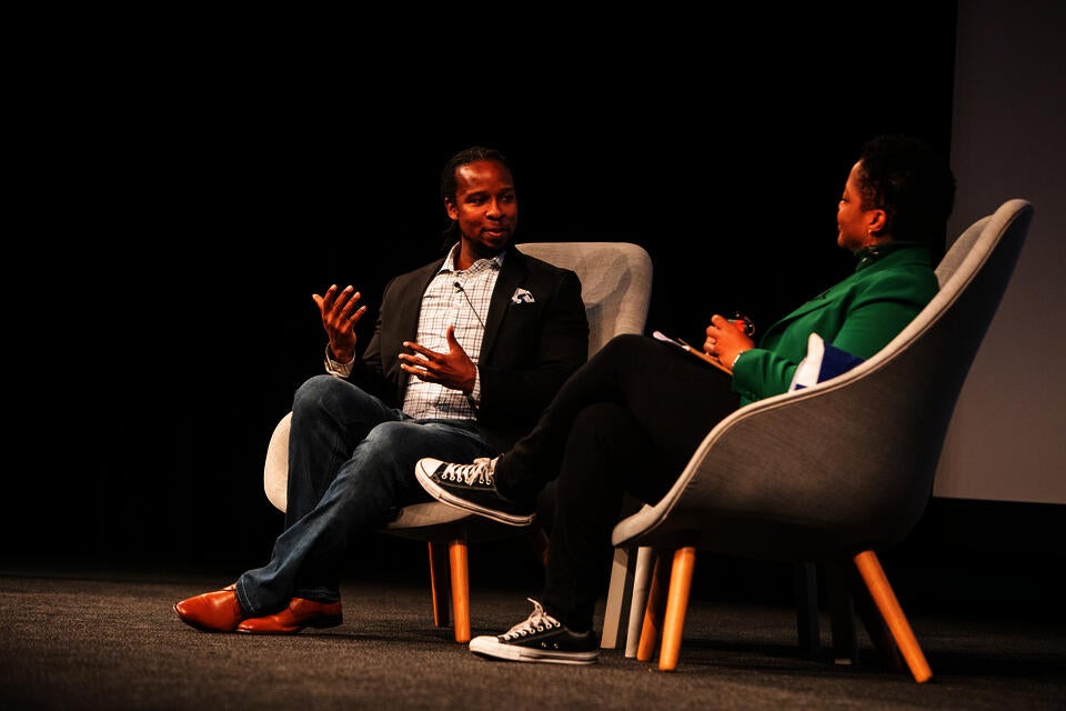 A Black man and woman sit on a stage. The man gestures and speaks as the woman looks on.