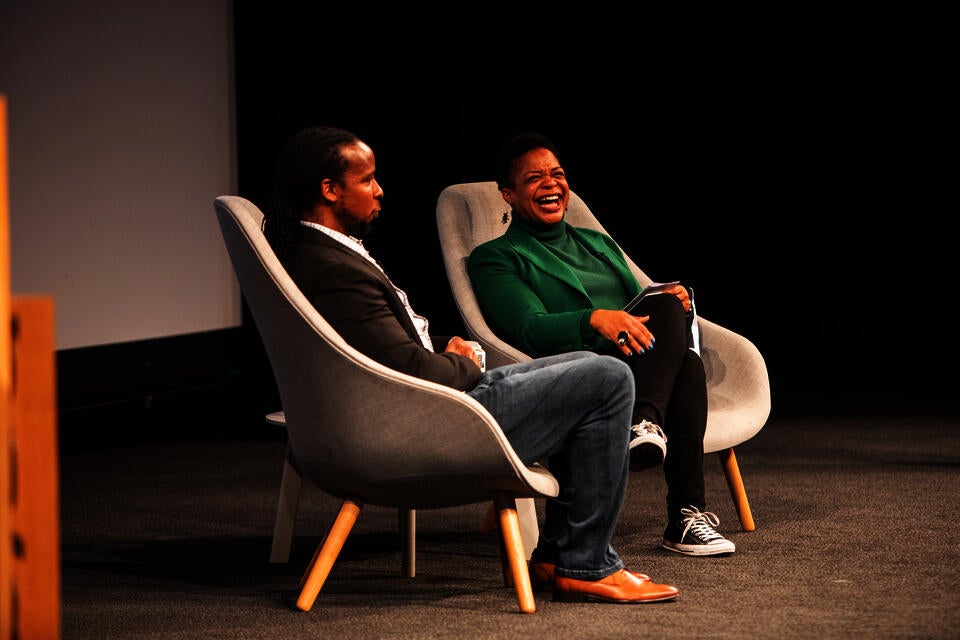 A Black man and woman sit on a stage. The woman, in a green blazer and turtleneck and black converse, laughs happily.