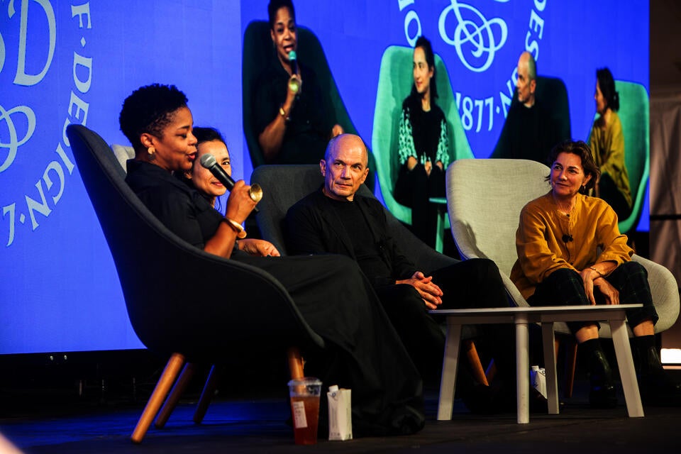 Three women and one man sit in chairs on a stage. One of them, a Black woman, speaks into a microphone. They are projected onto a screen behind them.