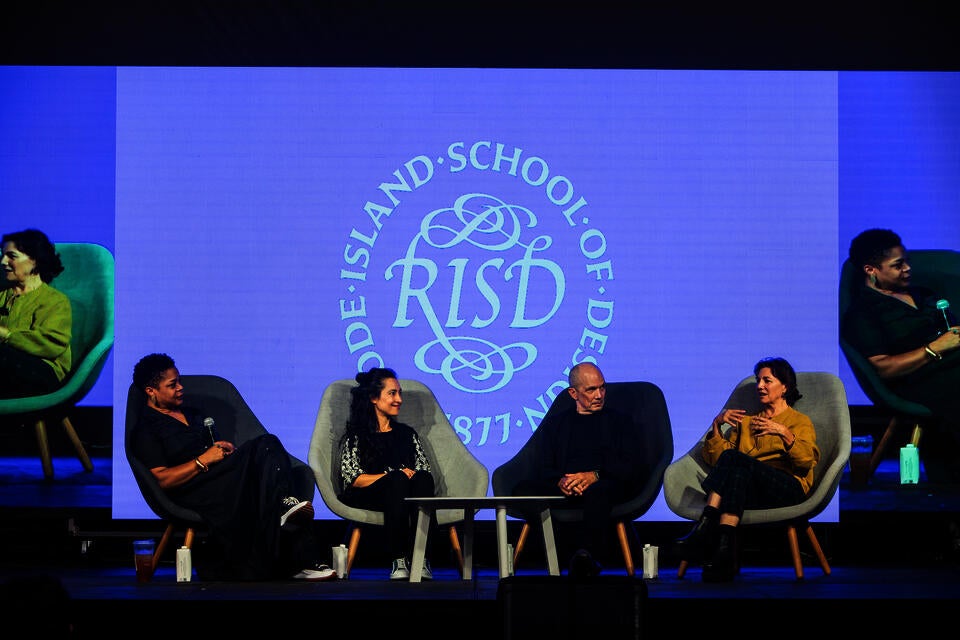 Four people sit in chairs on a stage. A white woman in a yellow sweater speaks while the others look on. The RISD seal is projected behind them.