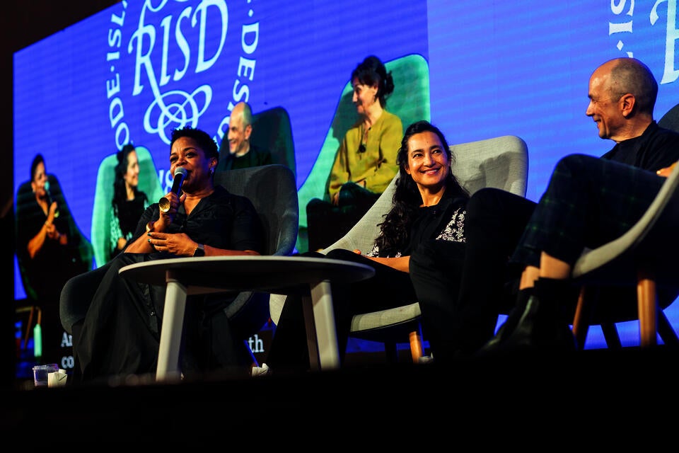 Low-angle photograph of two women and a man sitting on a stage. The Black woman speaks into a microphone.