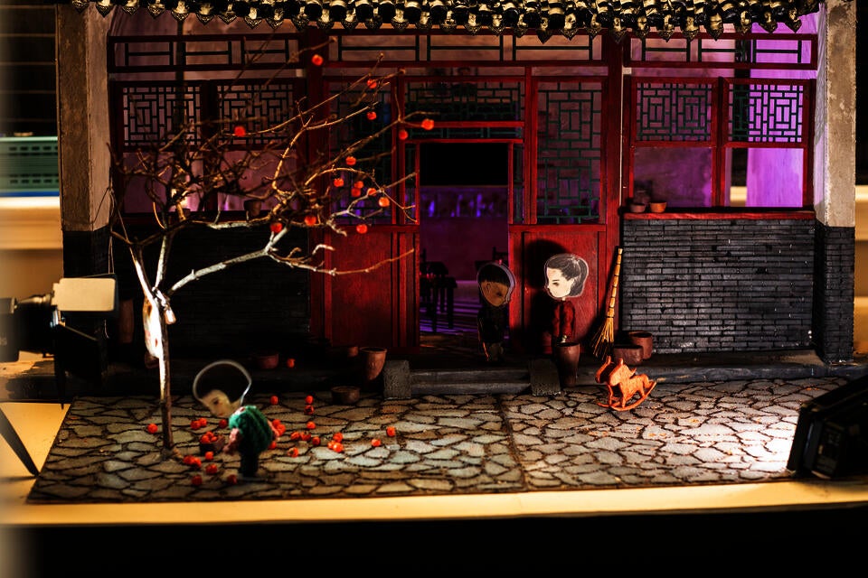 Small diorama set of a scene outside a house with Asian style architecture. In the foreground, and young girl figure picks up persimmons. 