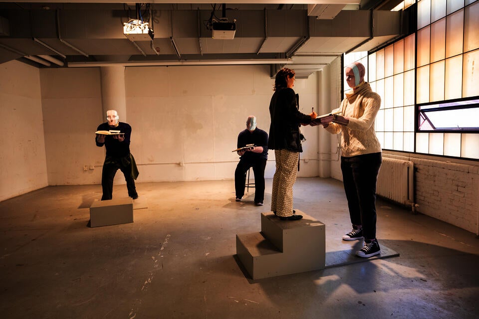 White gallery space with three robotic-looking human-sized figures dressed in regular clothes, holding books. A real human woman writes in one of their books. 