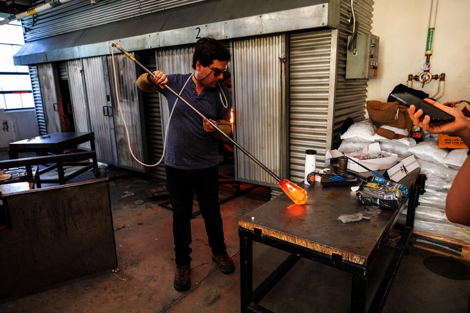 A man demonstrates blowing glass. He holds a pipe with molten glass on the end, rolling it on a table as he blows into a tube connected to the pipe.