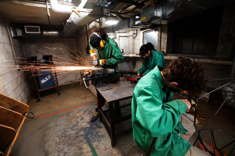 Three students work in a metals shop wearing green protective smocks. Sparks fly as one student cuts through metal.