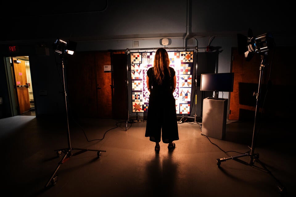 A woman stands in a classroom, in front of a suspended quilt lit by two powerful fresnel lights with barn doors.
