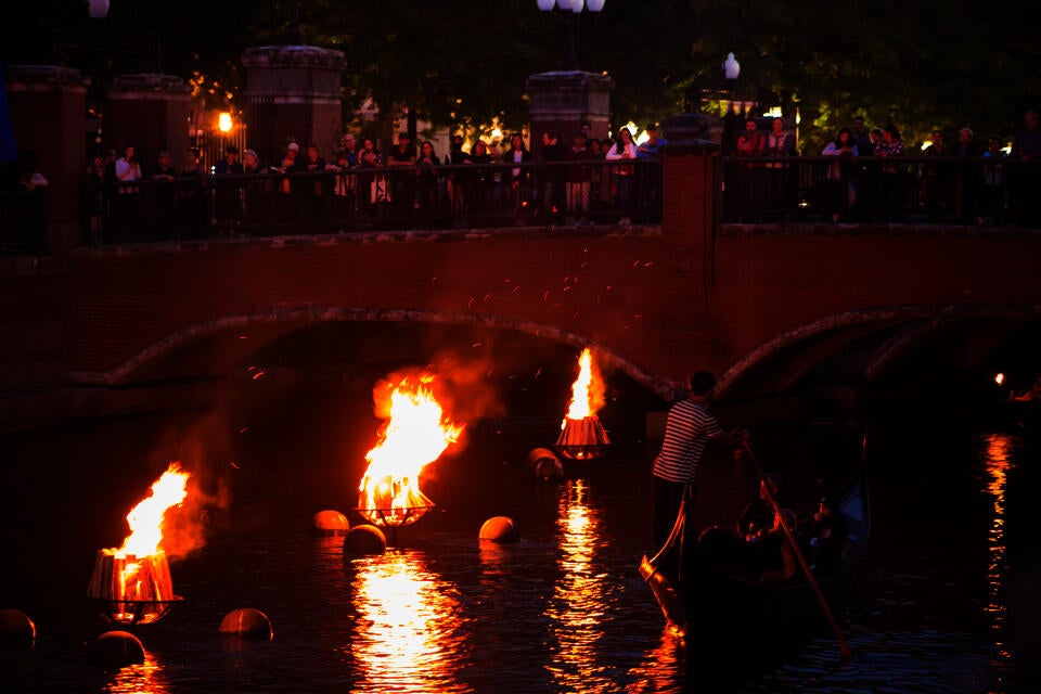 Providence WaterFire. At night, a crowd gathers on the College Street bridge overlooking the Providence River. The river is lit by burning braziers. A man rows a gondola under the bridge.