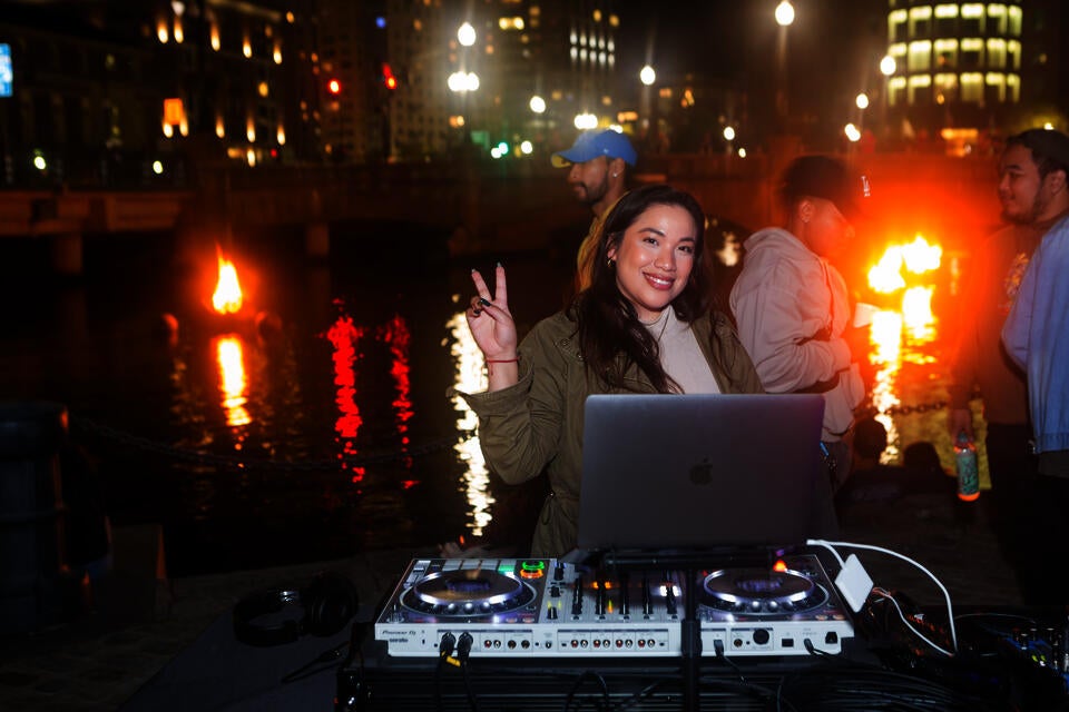 At night, a smiling young woman stands in front of DJ equipment, raising her hand in a peace symbol. She has long green nails. In the background, braziers burn in the Providence river.