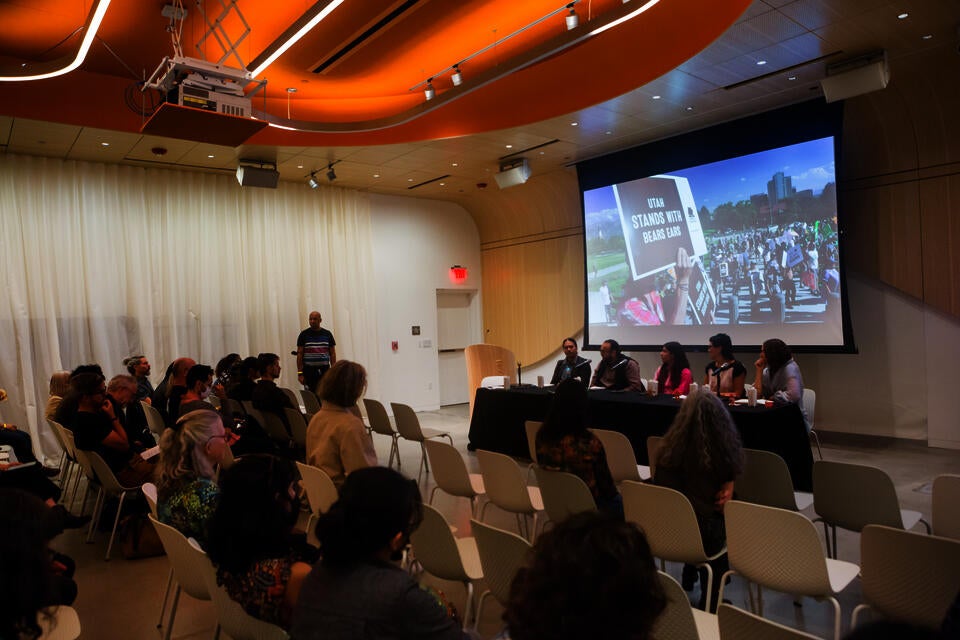 A panel of five people present in front of a seated audience. A projection behind them shows a peaceful protest with someone holding a sign that says 'Utah stands with Bears Ears.'
