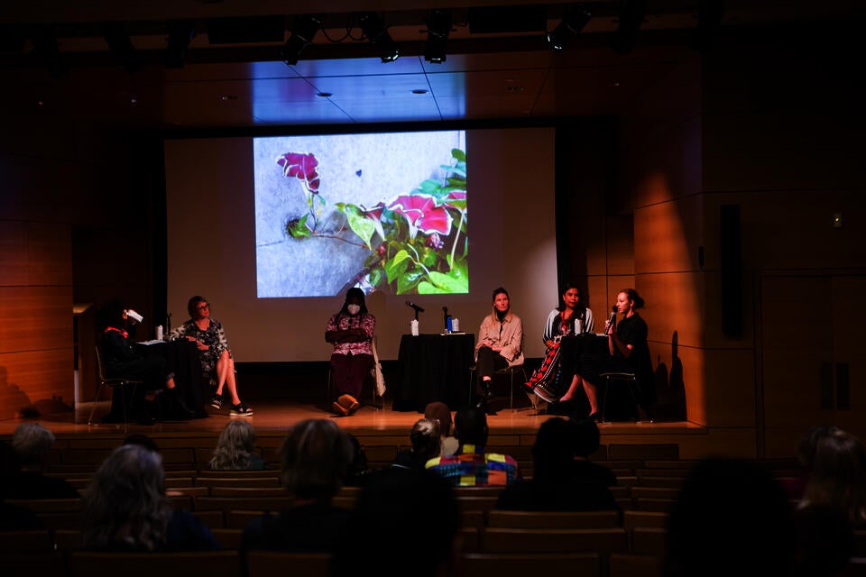 Four panelists and two moderators sit on stage at a wood-paneled auditorium. An image of a flowering vine is projected behind them.