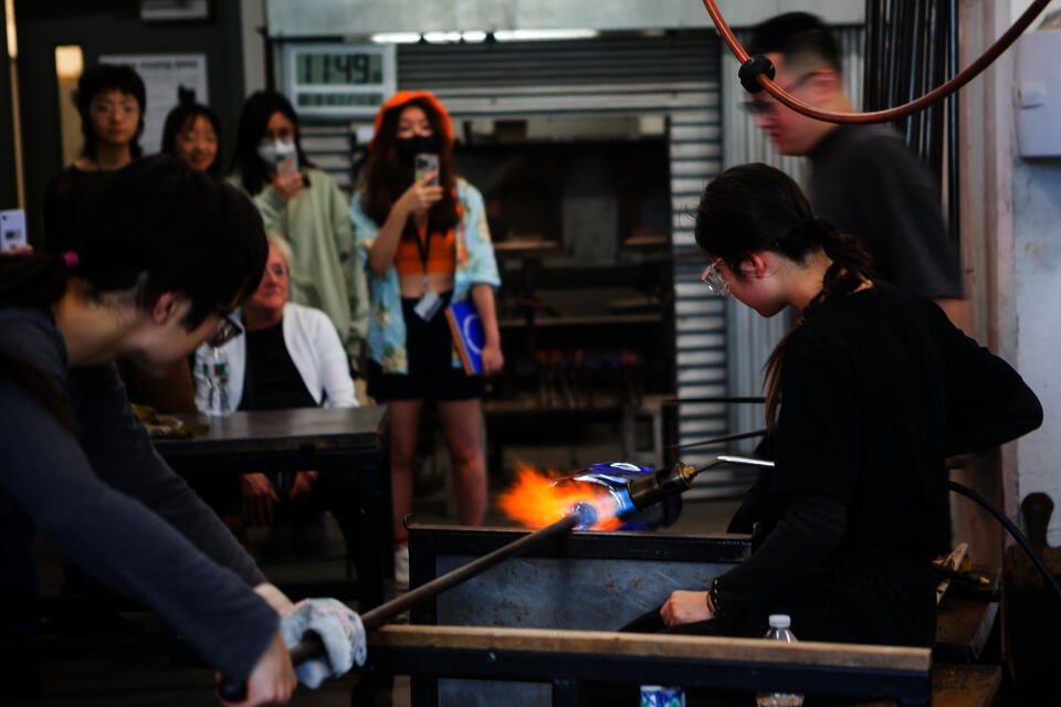 Two students demonstrate blowing glass, using a torch to heat glass on a long pipe,  while others look on, recording with their cellphones.