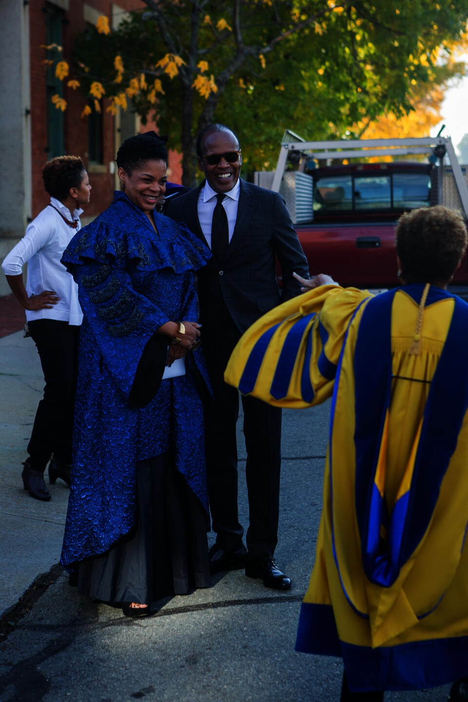 A Black woman in blue ceremonial robes and a Black man in a suit greet a woman in yellow and blue doctoral robes