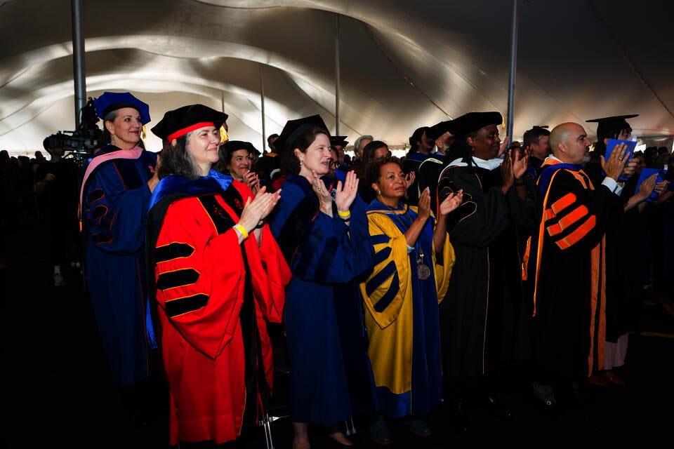 A group of adults in doctoral robes of various colors stand and clap in a tent