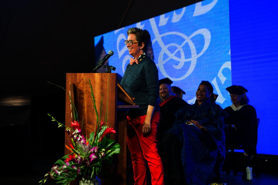 A white person in red pants stands at a podium on a stage, with a group of seated people in doctoral rows looking on
