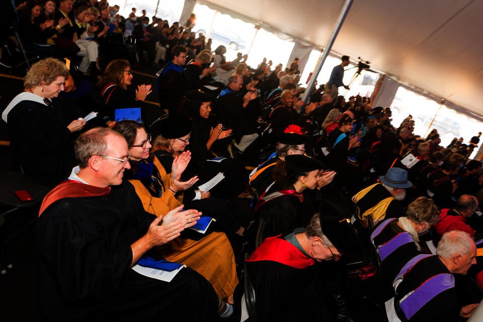 A crowd of seated people dressed in masters and doctoral robes applaud.