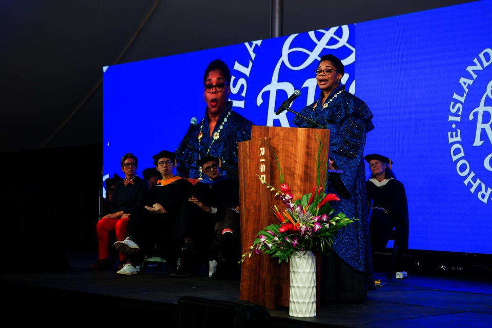 A Black woman in blue ceremonial robes speaks at a podium, on a stage where a group of onlookers sit. Her image is projected behind them.