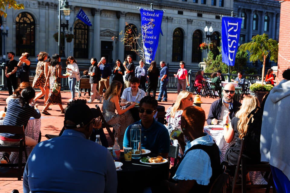 On a sunny Fall day, groups sit and eat at small round tables. In the background, people walk along the Providence Riverwalk. Blue RISD banners hang from light poles.