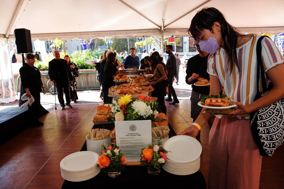 Buffet line outdoors under a tent. A table is piled with sandwiches, plates, and flowers. A young AAPI person picks up a plate in the foreground, while a small group examines food offerings in the background.