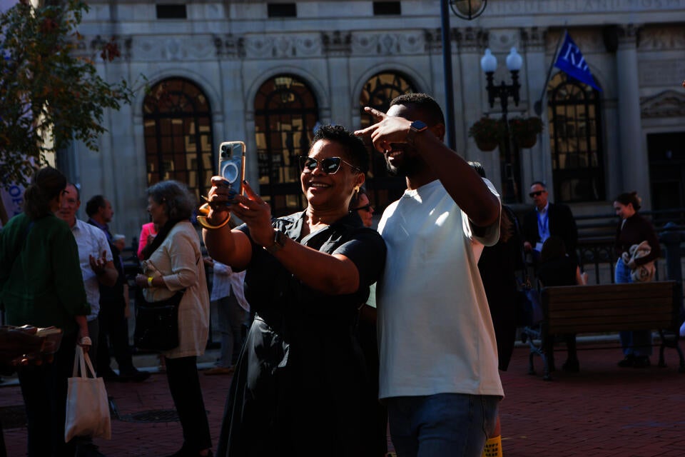 Outside on a brick walkway a Black woman in sunglasses takes a selfie with a young black man in a white tee shirt.