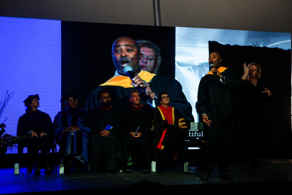 A black man in masters robes sings on a stage, with five professors seated, looking on. A woman stands behind him, interpreting in sign language.
