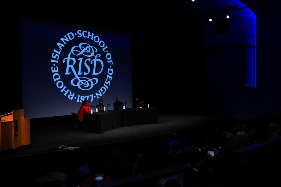 Five panelists sit at a table onstage in an auditorium. The RISD seal is projected behind them.