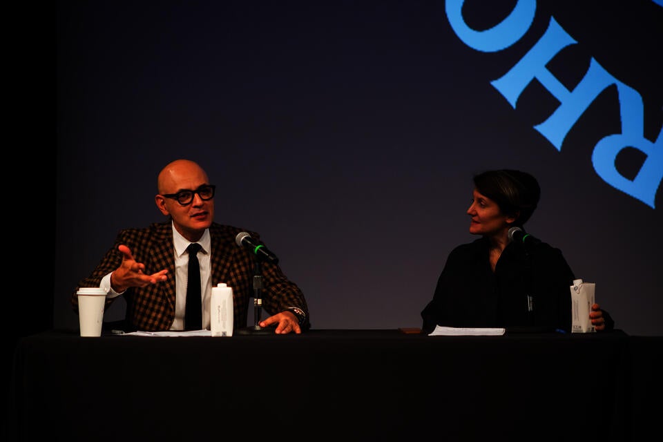 A nam in a yellow and black checked suit jacket gestures and speaks into a microphone. A woman in black on the right looks on, listening.