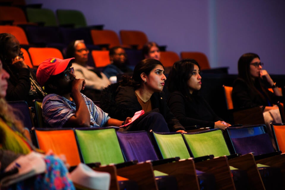 Interested observers sit and ponder in colorful auditorium chairs.