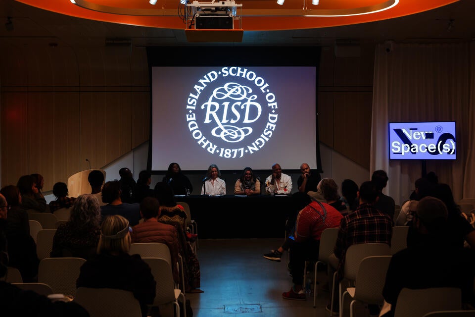 Five panelists and one moderator sit at a table in front of a small seated crowd. The RISD seal is projected behind them.