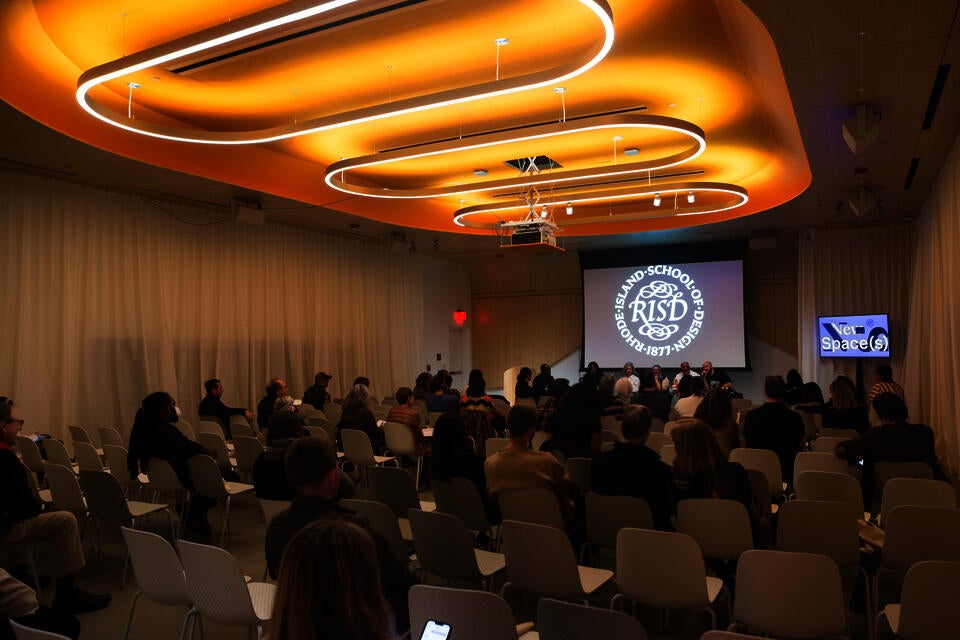 Five panelists and one moderator sit at a table with the RISD seal projected behind them. Large circular light fixtures hang above them.