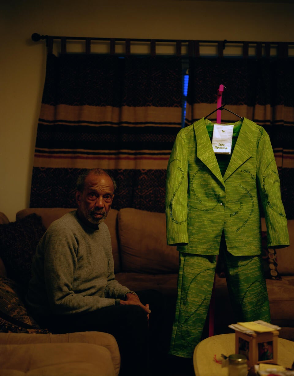 Man sitting in his living room on a couch with a green-colored suit hanging on a clothing rack to his right.