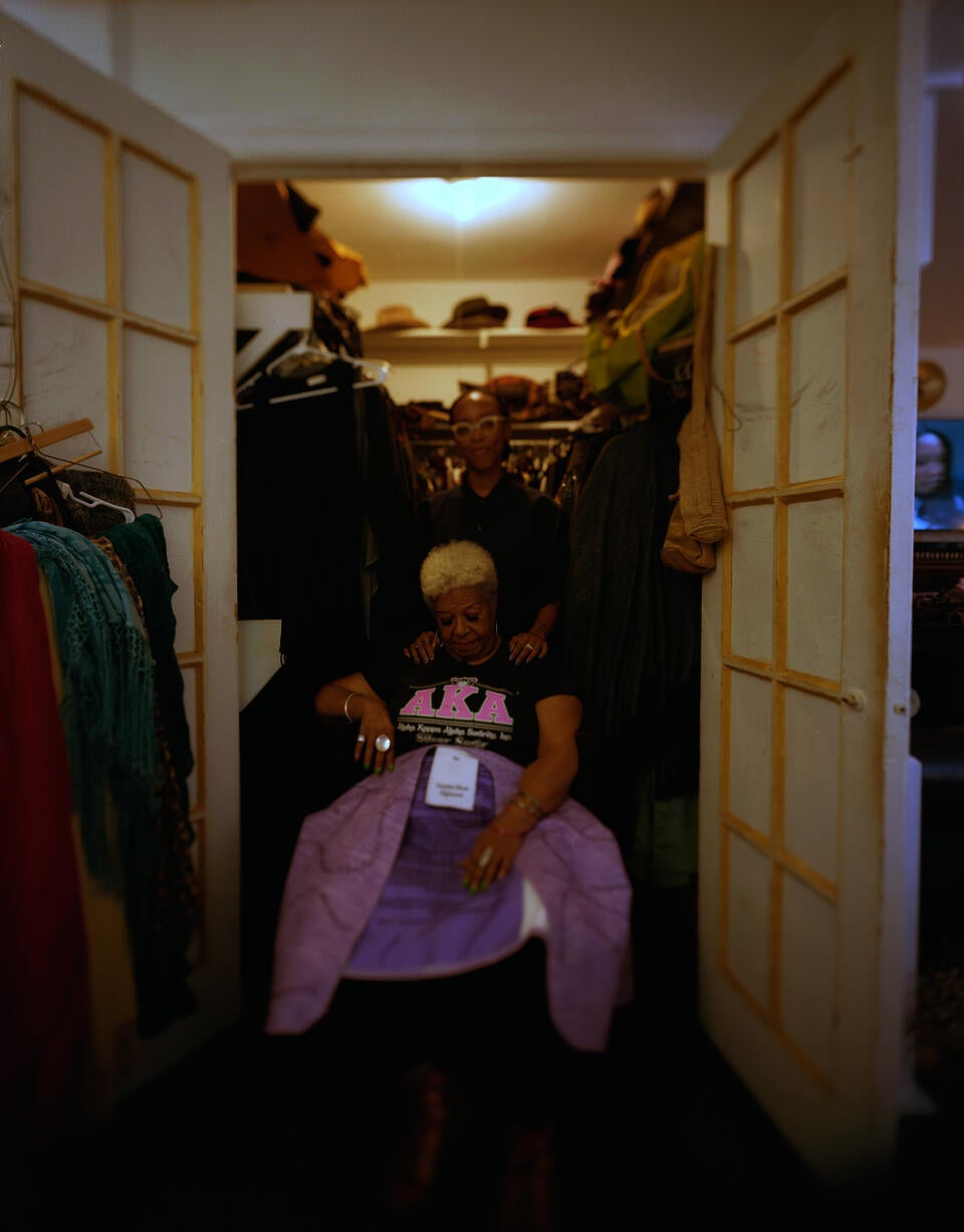 Woman sitting on a chair at the entrance of a double-door closet. She is holding a purple dress and looking down at it upon her lap. Behind her stands a woman smiling and looking at the camera.