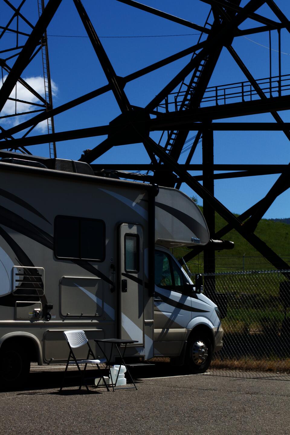 A camper van parked in front of the Mountain Con headframe. The swishy pattern of the camper's exterior seems to mirror the steel that criss-crosses the blue sky. The door of the camper is slightly ajar, and a camping chair and table sit on the parking lot outside the vehicle.. 