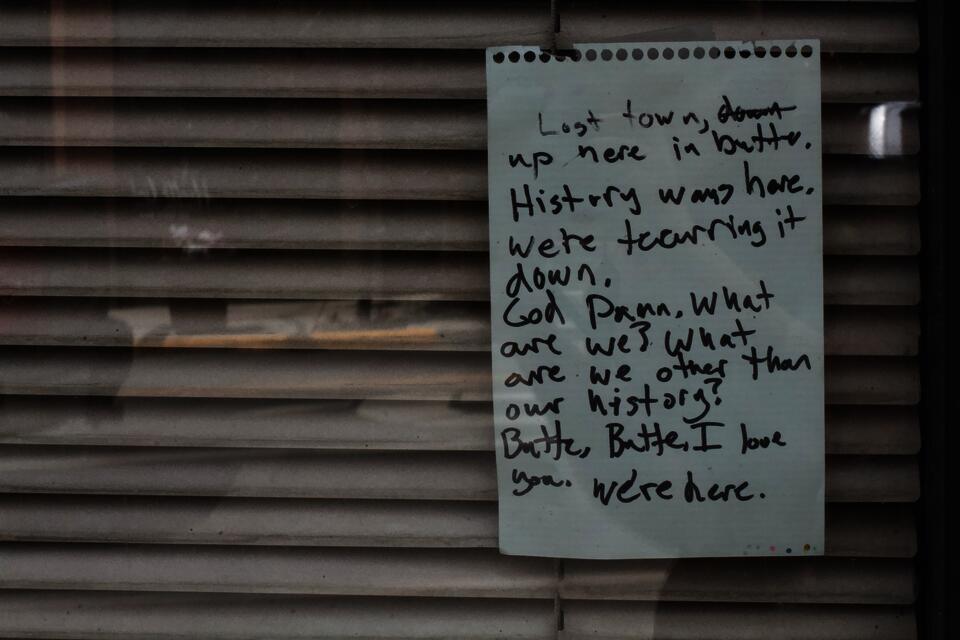 A note taped inside of a bar window that reads, "Lost town, [down is crossed out] up here in Butte. History was here. We're tearing it down. God damn, what are we? What are we other than our history? Butte, Butte, I love you. We're here." The blinds inside the bar window are shuttered.