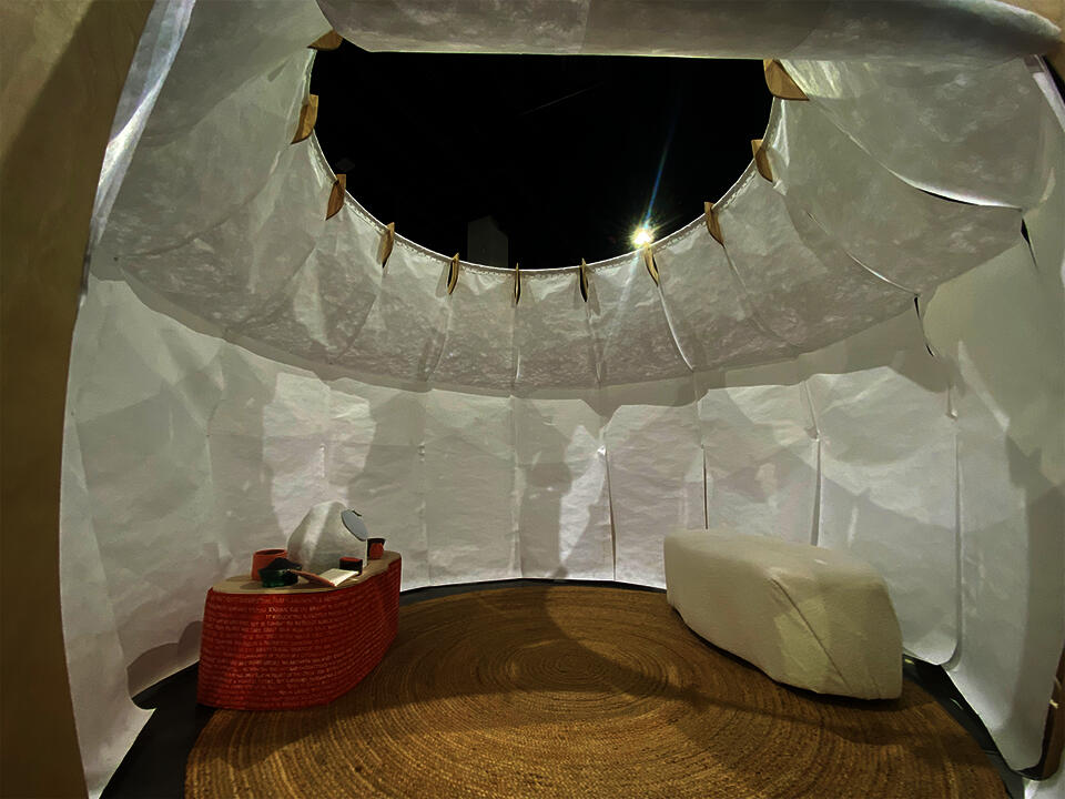 The interior of a white stand-alone structure houses an orange table with a white ceramic speaker, dried herbs, and cards.