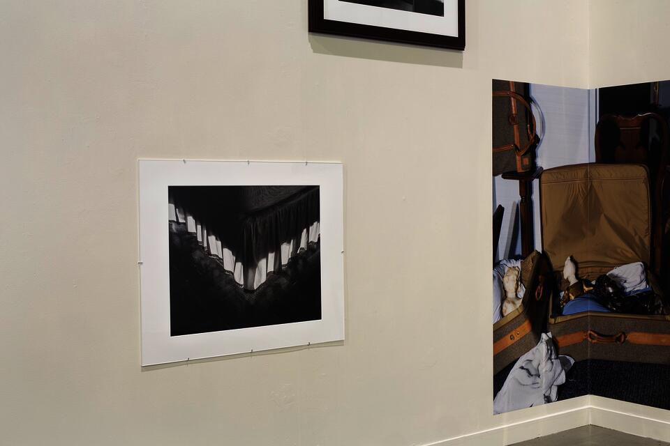 Arrangement of three photographs, one of a bed skirt mounted under a mat board and plexiglass, another color pigment print mounted in a corner, the third barely visible above.