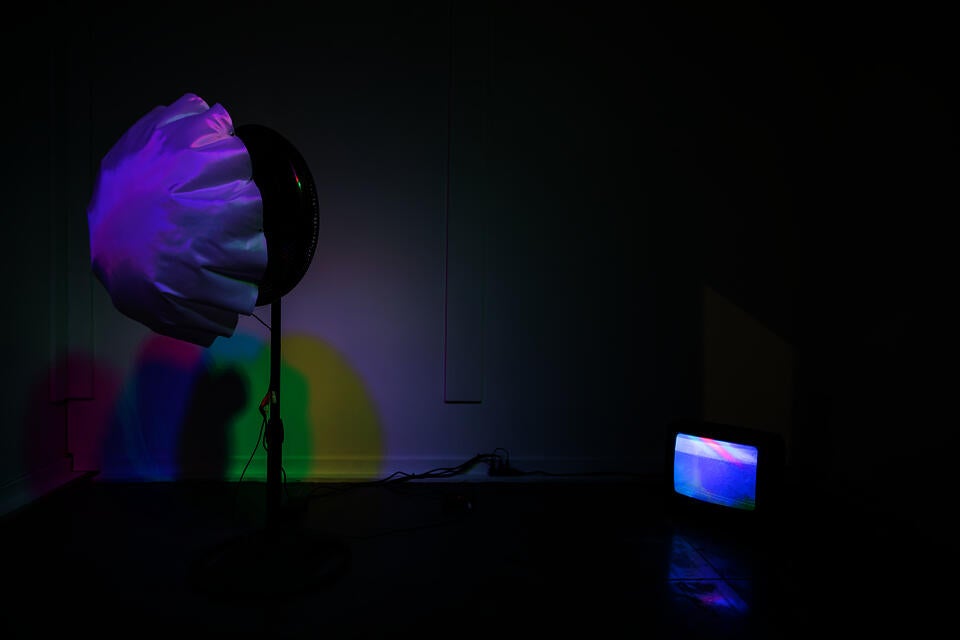 White fabric is attached to a fan and being blown up under colorful light.