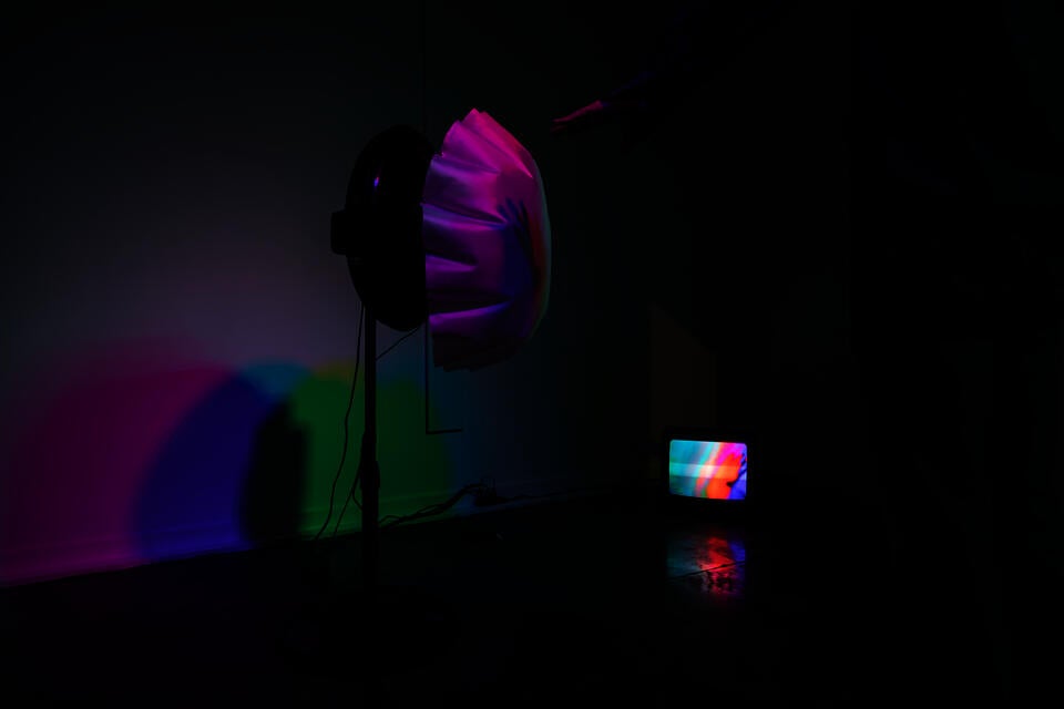 White fabric is attached to a fan and being blown up under colorful light.