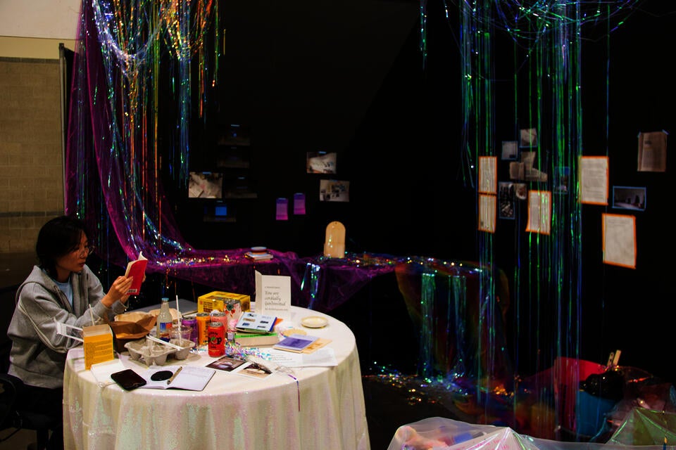 books, food, and drinks on white table; iridescent party streamers hanging from above like curtain; photos and writings on corner-shaped black walls