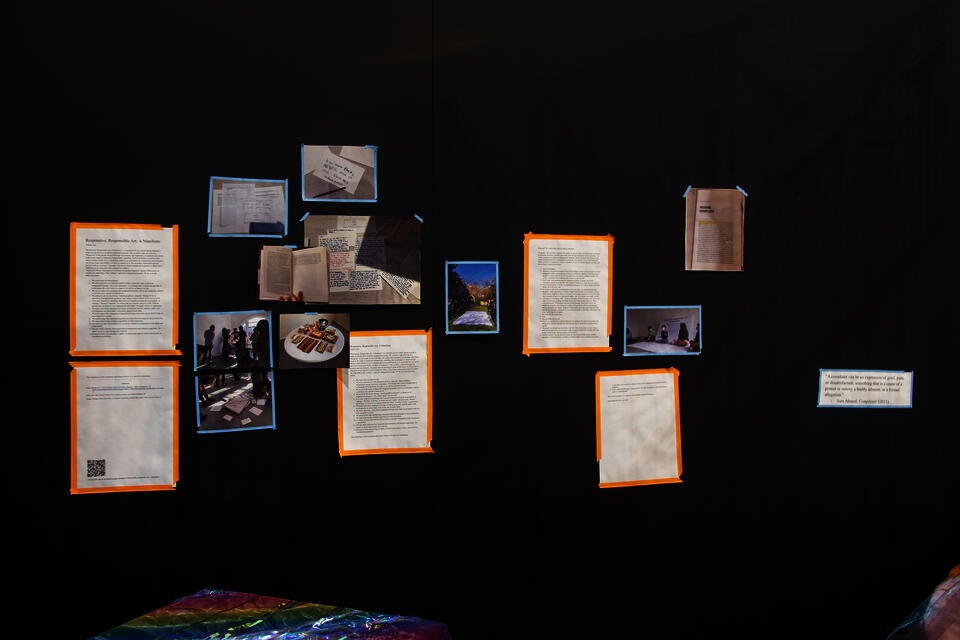 books, food, and drinks on white table; iridescent party streamers hanging from above like curtain; photos and writings on corner-shaped black walls