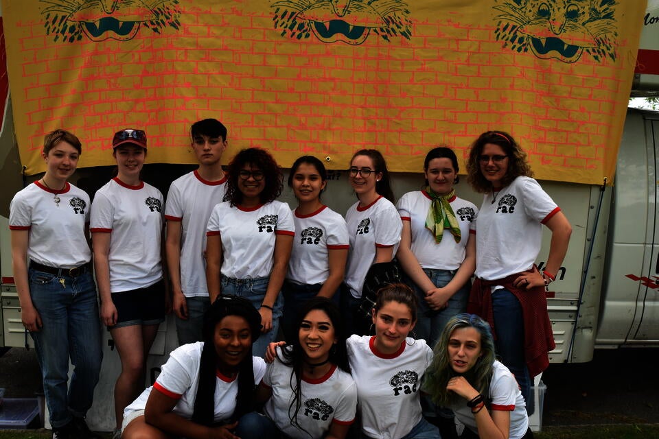 Twelve teenagers in two rows all wearing a white t-shirts with a maroon collar in front of beige fabric banner with blue monsters. 