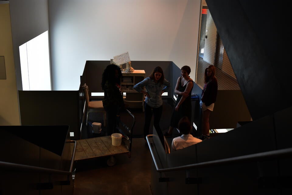 Camera angled from the top of the stair looking down a staircase at five people talking  with some art installation equipment nearby.