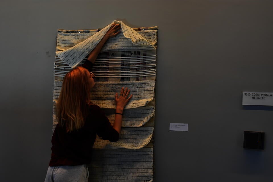 Student straightening a wall piece that has layers of fabric one of which is being lifted to reveal small letters that spell Call Mom.