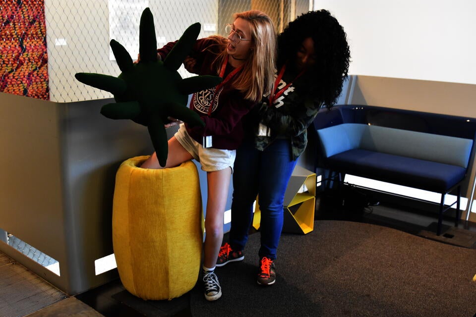 Teenage helping another teenager step into a two foot pinneapple shaped stool that is holding the leaf shaped stool top. 