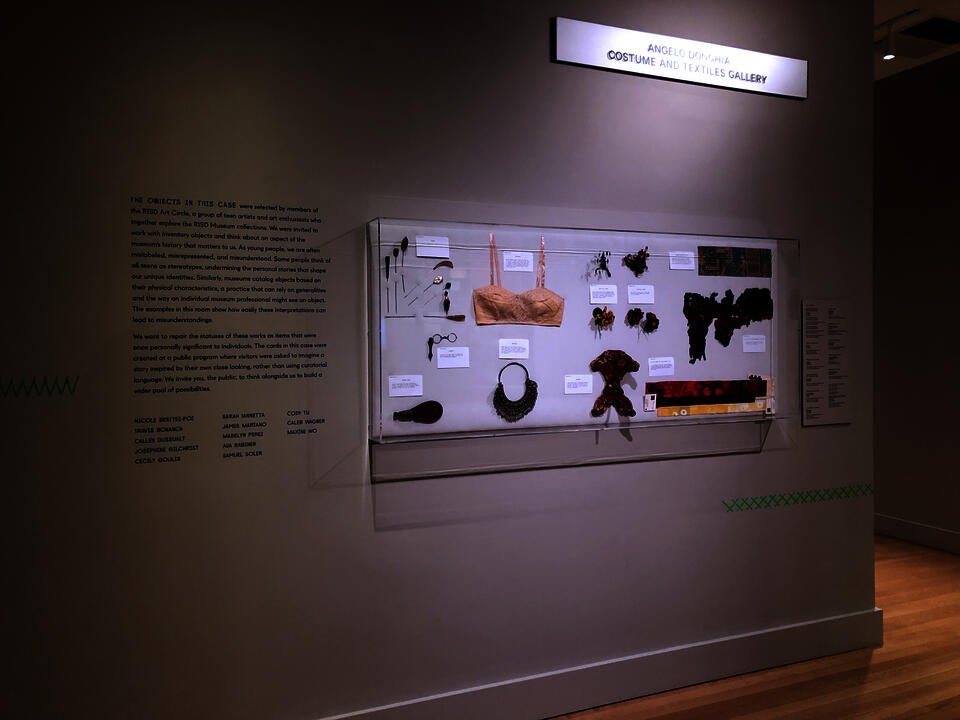 A case on a wall with variety of object including a pieces fabric both ancient and modern, chinese hat stand, a bra, costume jewelry, hand-made fake flowers, and hat pins