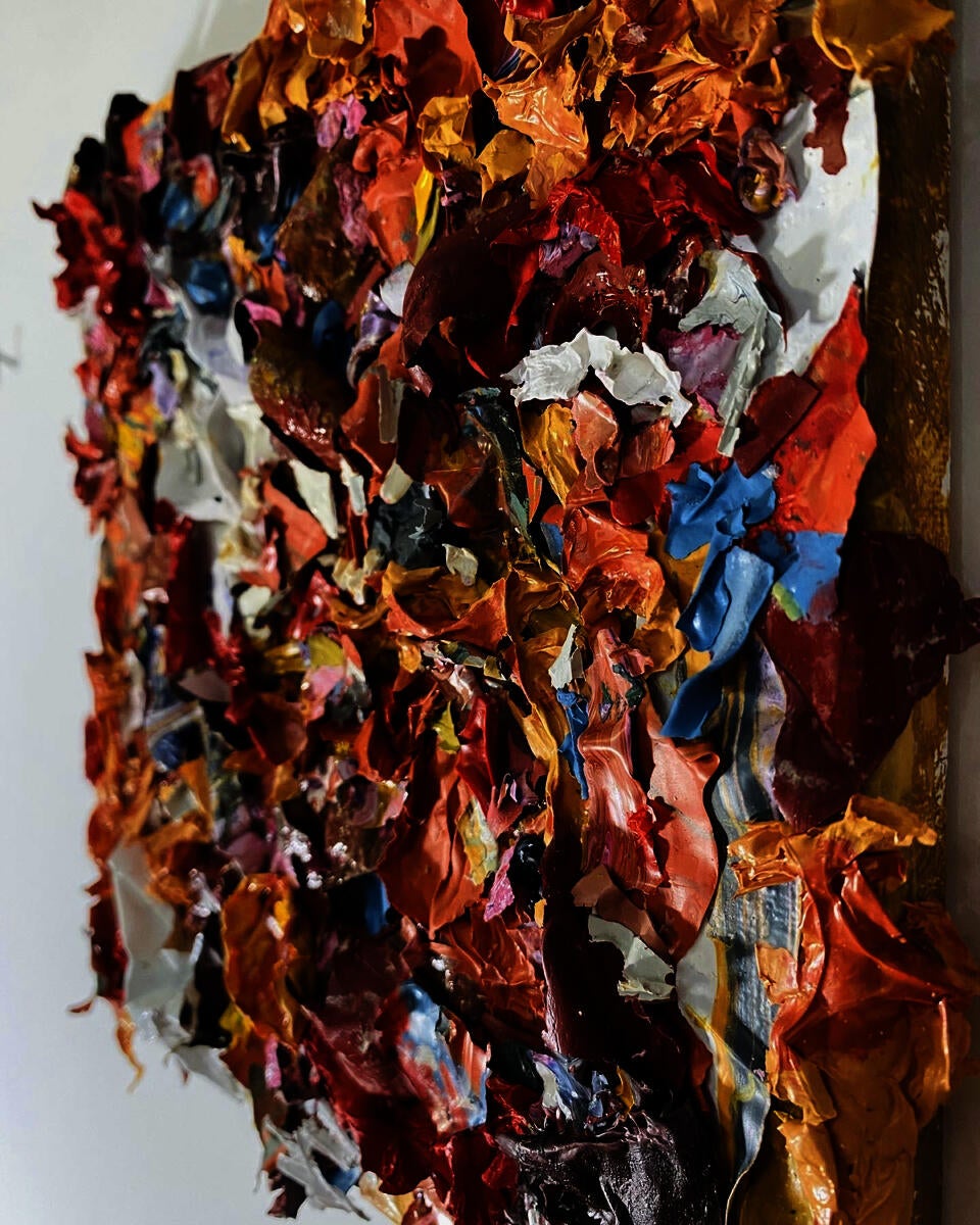 Small pieces of scrap paint are glued onto a canvas. The pieces vary but are roughly 1"x1". The scrap colors are red, yellow, blue, orange and white.